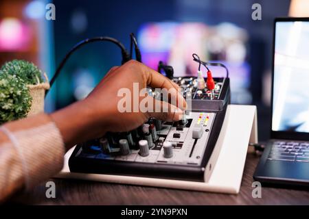 Nahaufnahme einer afroamerikanischen Person, die den Sound-Mixer für gute Qualität bei der Aufnahme von Podcast-Episoden optimiert. Selektiver Fokus des schwarzen individuellen Einstellknopfs der Audioanlage während der Talkshow. Stockfoto