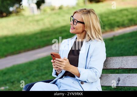 Eine fröhliche, lächelnde Profi-Frau, die ihr Smartphone beiläufig benutzt, während sie im Park sitzt Stockfoto