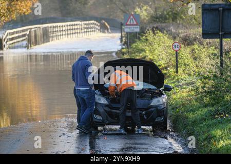 Lacock, Wiltshire, Großbritannien. November 2024. Der starke Regen des Sturms Bert hat den Fluss Avon in Lacock überschwemmt. Die Straße ist für alle bis auf die größten Fahrzeuge unpassierbar. Einige Fahrzeuge schafften es nicht durch die Flut. Dieser Standort wird mehrmals im Jahr überschwemmt, aber heute ist das Wasser viel höher, und das Hochwasser erstreckt sich normalerweise nicht über den Damm hinaus, sondern hat es heute getan. Männer reparieren ein Auto, das bei der Flut kaputt ist Stockfoto