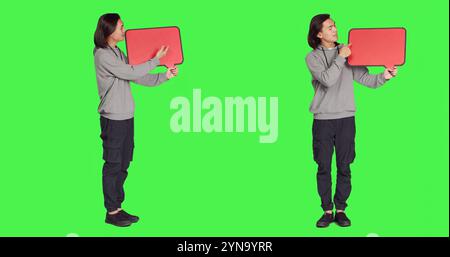 Männliches Modell posiert mit Sprechblase und präsentiert leere Pappe mit Copyspace vor dem Ganzkörper-Greenscreen. Asiatische Person, die rote Kartonplatte für Marketingwerbung hält. Stockfoto