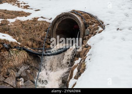 Wasser fließt aus der Sturm-Entwässerungsleitung, umgeben von schmelzendem Schnee auf einer felsigen Landschaft. Stockfoto