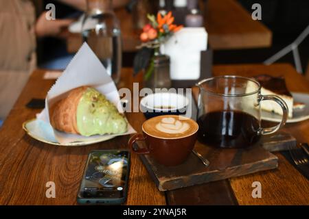 Ein entzückendes Café mit einem Latte mit Kunst, einer Teekanne Kaffee, einem Buttercroissant und einem Käsekuchen. Absolut gemütlich Stockfoto