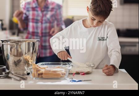 Ein kleiner Junge mischt Creme in einer Schüssel auf einer Küchentheke, während ein Erwachsener im Hintergrund zu sehen ist. Verschiedene Backwerkzeuge und ein Dessertgericht sind in der Nähe, ich Stockfoto