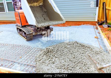 Schwere Maschinen geben frischen Beton auf vorbereitete Flächen ab. Neue Terrassenarbeiter führen den Prozess im Vorort mit einem kompakten Raupenkipper Stockfoto