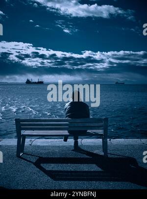 Eine Silhouette eines Mannes, der allein auf der Bank sitzt und auf das Meer blickt Stockfoto