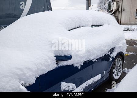 Schneebedecktes, typisches blaues Fließheck-Auto, das nach starkem Schneefall auf der Straße geparkt wurde. Weitwinkelansicht aus der Nähe, keine Personen. Stockfoto