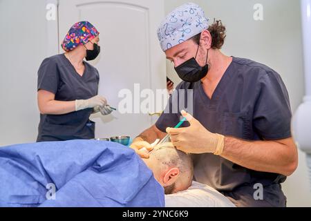 Ein Mann mit weißem Hut und schwarzem Mantel führt eine Operation an einem Glatzen durch. Der kahlköpfige Mann trägt ein blaues OP-Kleid Stockfoto