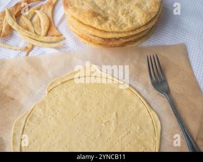 Die Rohschicht wurde auf einem Backpapier ausgerollt und mit einer Gabel, einem Haufen gebackener dünner Butterschichten und Teigresten auf einem weißen Waffeltuch durchbohrt. Stockfoto