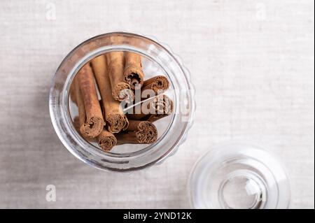 Ceylon Zimtfedern in einem Glasgefäß mit Deckel, auf Leinengewebe, mit leerem Platz für Text. Ganze, getrocknete Rindenstreifen von Cinnamomum verum. Stockfoto