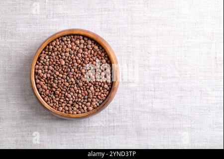 Getrocknete braune Linsen in einer Holzschale auf Leinengewebe, mit leerem Platz für Text. Ungeschälte Berglinsen mit braunen Samenmänteln und orange-roten Samen. Stockfoto