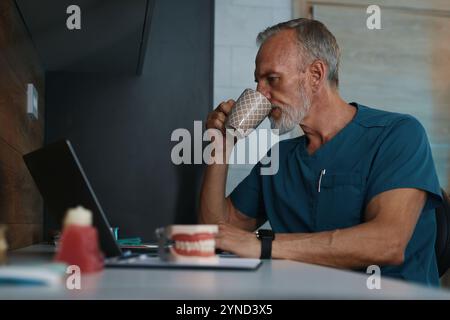 Ein älterer Arzt schlürft Kaffee am Schreibtisch, ohne in die Kamera zu schauen Stockfoto
