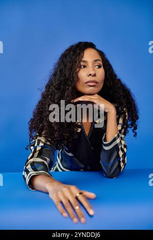 Eine schöne Frau legt ihr Kinn auf ihre Hand und zeigt ihre atemberaubenden Locken und stilvolle Kleidung. Stockfoto