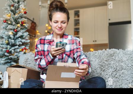 Frau, die zu Hause auf einem Sofa sitzt und während der Weihnachtsfeiertage Internetbestellungen auspackt und Tags ihrer Online-Einkäufe scannt. Stockfoto