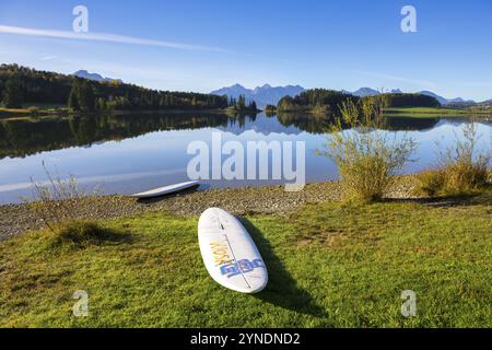 Ein Surfbrett liegt am Ufer eines ruhigen Sees mit Bergblick und Herbstlandschaft unter klarem Himmel, Forggensee bei Füssen, Ostallgäu, Bavari Stockfoto