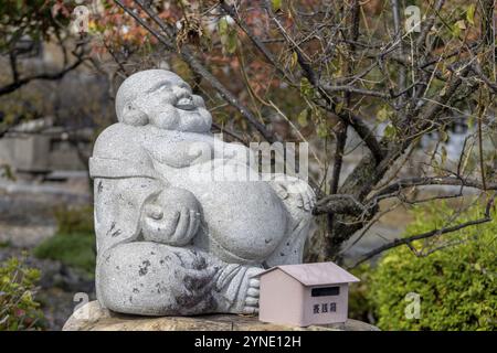 Nagahama, Präfektur Shiga, Japan. November 2023. Eine sitzende, lächelnde Buddha-Steinfigur an einem japanischen Shinto-Schrein Stockfoto