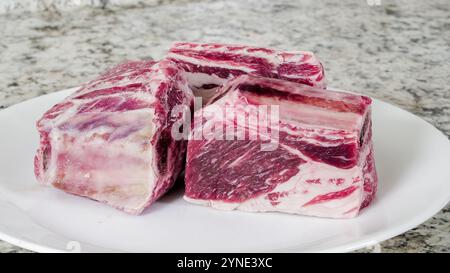 Drei große Stücke Rindfleisch-Rippchen werden ordentlich auf einem weißen Teller auf einer strukturierten Küchenarbeitsplatte präsentiert. Stockfoto