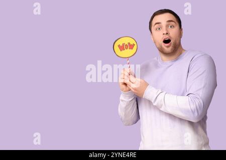 Schockierter junger Mann, der eine Sprechblase mit Wort WOW auf Fliederhintergrund hält Stockfoto