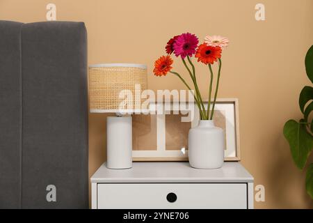 Vase mit Gerbera-Blumen, Bilderrahmen und Lampe auf Kommode in der Nähe der beigefarbenen Wand im Zimmer Stockfoto
