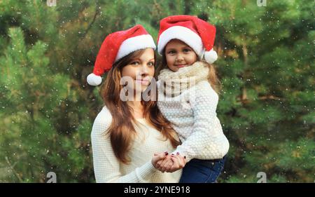 Familie, Feier, Urlaub. Glückliche Mutter umarmt ihr kleines Mädchen mit weihnachtsmütze zusammen in der Nähe des Weihnachtsbaums im Wald mit Schnee Stockfoto