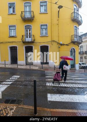 Ältere Frau, die an einem regnerischen Tag die Straße mit einem roten Regenschirm und Einkaufswagen entlang ging. Gelbes Gebäude im Hintergrund. Lissabon, Portugal Stockfoto