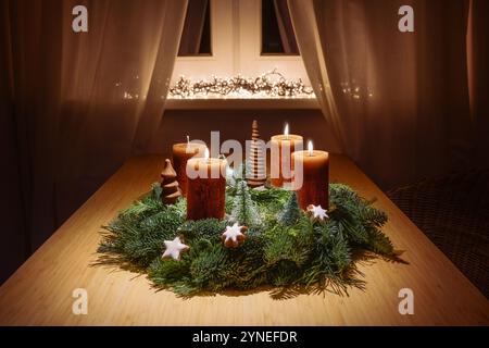 Dritte Adventszeit mit orangebraunen Kerzen auf einem Kranz aus grünen Tannenzweigen mit Weihnachtsdekoration auf einem Holztisch am Fenster in warmem Licht, Co Stockfoto