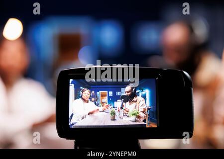 Konzentrieren Sie sich auf die professionelle Kamera, mit der Sie einen Live-Chat zwischen dem Moderator einer Talkshow und einem Mann im Heimstudio filmen. Nahaufnahme der Produktionsausrüstung mit Moderator und Gast, die in unscharfem Hintergrund chatten. Stockfoto