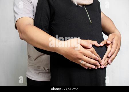 Ein Mann umarmt eine schwangere muslimische Frau von hinten, ohne erkannt zu werden. Konzept eines Schwangerschaftsfoto-Shootings mit weißem Hintergrund Stockfoto