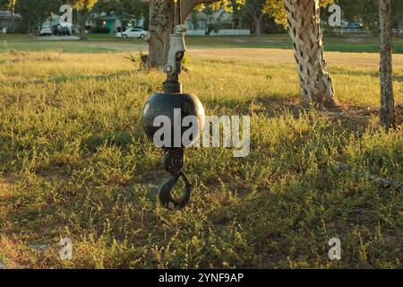 Ein isolierter schwarzer Krankugelhaken hängt an einem Kabel, das an einem Kran befestigt ist. Grünes Gras und Bäume im Hintergrund. Stockfoto