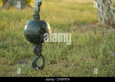 Ein isolierter schwarzer Krankugelhaken hängt an einem Kabel, das an einem Kran befestigt ist. Grünes Gras und Bäume im Hintergrund. Stockfoto