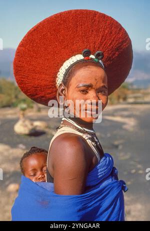 Porträt einer schönen Zulu-Frau, mit ihrem Baby, in traditioneller Kleidung und mit Isicholo-Hut. Stockfoto