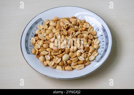 Geröstete Erdnüsse (Arachis hypogaea) auf einem weißen Teller auf einem hellen Holzschutt. Stockfoto