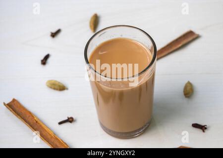 Ein Glas Masala Chai auf einer hellen Holzoberfläche. Verstreute Zimtstangen, Kardamom und Nelken verleihen der Szene einen Hauch von Würze. Stockfoto