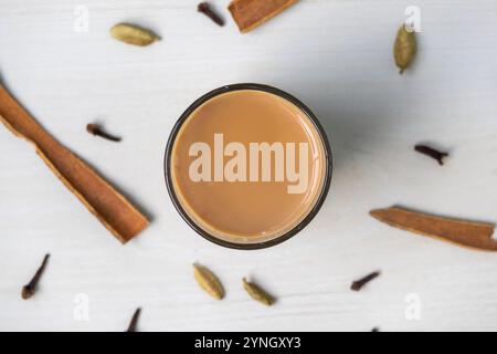 Blick von oben auf Masala Chai auf einer Glasschale auf einer hellen Holzoberfläche. Verstreute Zimtstangen, Kardamom und Nelken verleihen der Szene einen Hauch von Würze. Stockfoto