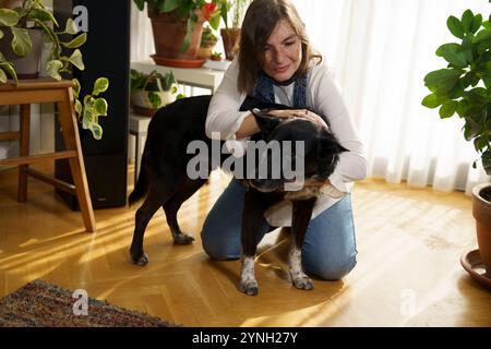 Eine Frau, die auf dem Boden kniet und sanft einen schwarzen Hund umarmt in einem sonnendurchfluteten Raum voller Pflanzen. Die Atmosphäre ist warm und gemütlich, mit sanften Schatten auf t Stockfoto