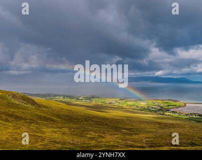 Ein lebhafter Regenbogen durchzieht den Himmel über einer üppig grünen Landschaft mit einem Ozean am Horizont. Die Szene wird von dramatischen Wolken beleuchtet Stockfoto