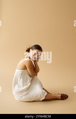 Eine junge Frau sitzt anmutig in einem Studio und zeigt ihr elegantes Kleid und ihr selbstbewusstes Verhalten. Stockfoto