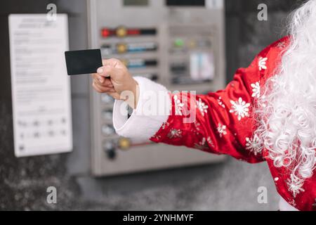 Nahaufnahme eines Weihnachtsmanns mit Kreditkarte in der Nähe eines Zahlungsterminals. Eine moderne Interpretation der Weihnachtsfigur mit Technologie in festlicher Umgebung. Stockfoto