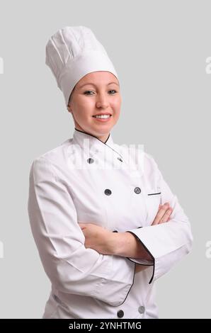 Junge Küchenchefin lächelt mit überkreuzten Armen, trägt weiße Uniform und eine Haube Stockfoto