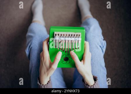 Frau, die eine grüne Kalimba spielt, ein traditionelles afrikanisches Musikinstrument, sitzt mit weißen Socken und Jeans auf dem Boden Stockfoto