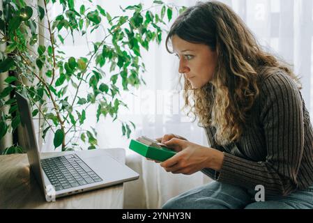 Junge Frau, die Kalimba spielt, nach einer Online-Musikstunde mit ihrem Laptop zu Hause, mit einer Ficus-Pflanze im Hintergrund Stockfoto