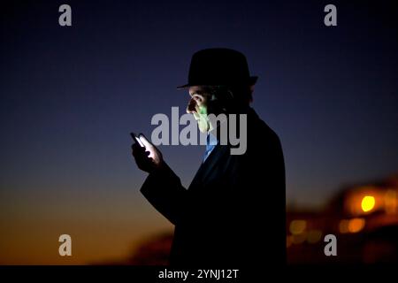 Ein Mann steht in der Silhouette vor dem Abenddämmerhimmel, konzentriert auf sein Smartphone. Er trägt einen Hut und befindet sich in der Nähe der Küste, mit fernem Licht Stockfoto