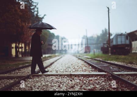 Ein Mann schlendert an einem regnerischen Tag entlang einer verlassenen Eisenbahnstrecke, während er einen Regenschirm hält. Der Hintergrund zeigt alte Zugwagen und Bäume, Kreatin Stockfoto