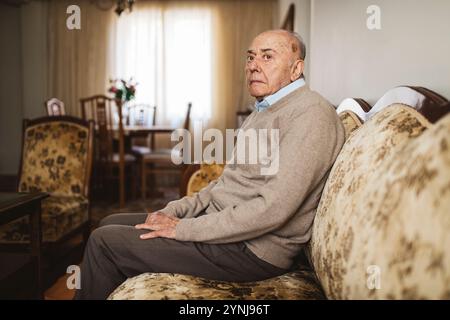 Ein älterer Mann sitzt auf einem Sofa mit Blumenmuster, trägt einen grauen Pullover und sieht nachdenklich aus. Sonnenlicht filtert durch Vorhänge und beleuchtet den Stockfoto