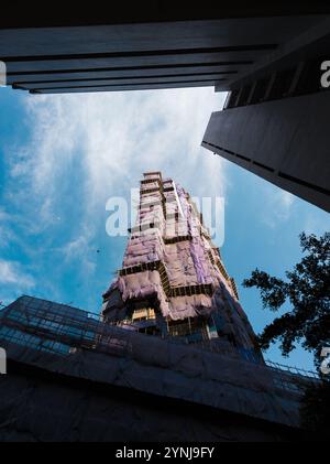 „Wolkenkratzer im Bau, umhüllt in lila Gerüste, aus einem niedrigen Winkel vor einem blauen Himmel mit gerahmten städtischen Strukturen“ Stockfoto