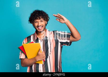 Foto von funky cool Guy gekleidetes gestreiftes Hemd, das Copybooks hält, Arm winkend hoch hoch gelegener Raum isolierter blauer Farbhintergrund Stockfoto