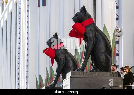 London, Großbritannien. 26. November 2024. Zwei große Katzenskulpturen, die den Eingang zum Greater London House in Mornington Crescent bewachen, sind mit festlichen roten Bögen für die Weihnachtszeit geschmückt. Das Art-Deco-Gebäude, ursprünglich die Carreras Zigarettenfabrik, ist ein Beispiel für die ägyptische Wiedergeburt-Architektur des 20. Jahrhunderts und die Katzen repräsentieren die ägyptische Göttin Bastet. Quelle: Ron Fassbender/Alamy Live News Stockfoto