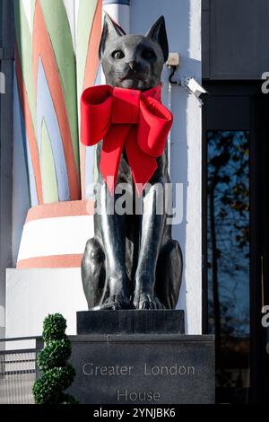 London, Großbritannien. 26. November 2024. Zwei große Katzenskulpturen, die den Eingang zum Greater London House in Mornington Crescent bewachen, sind mit festlichen roten Bögen für die Weihnachtszeit geschmückt. Das Art-Deco-Gebäude, ursprünglich die Carreras Zigarettenfabrik, ist ein Beispiel für die ägyptische Wiedergeburt-Architektur des 20. Jahrhunderts und die Katzen repräsentieren die ägyptische Göttin Bastet. Quelle: Ron Fassbender/Alamy Live News Stockfoto