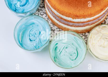 Farbenfrohe Zuckerguss in Glasschüsseln, die frisch gebackenen Schichtkuchen auf dekorativem Teller umgeben Stockfoto