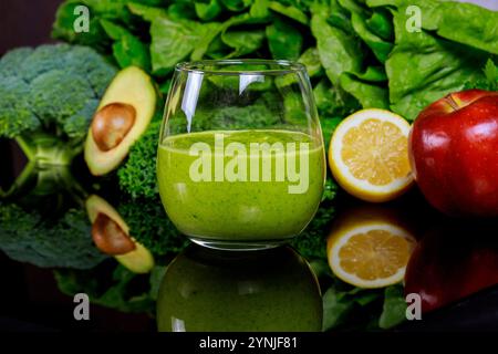 Frischer grüner Smoothie mit Avocado, Apfel und Zitrone auf reflektierender Oberfläche, umgeben von Grüntönen Stockfoto