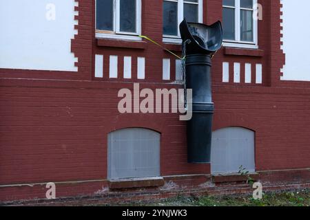 Ein markanter schwarzer downspout ist an einem roten Backsteingebäude angebracht, der sich nach unten zum Boden erstreckt. Unkonventionelle Platzierung lenkt die Aufmerksamkeit auf den Stockfoto